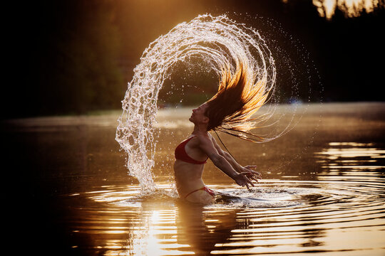 Woman in red bikini flipping hair in arc of water at sunset, creating a dynamic splash. WA, USA