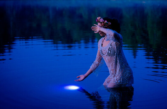 Woman in lace dress kneeling in a lake at night, hand shielding from mystical blue light. WA, USA