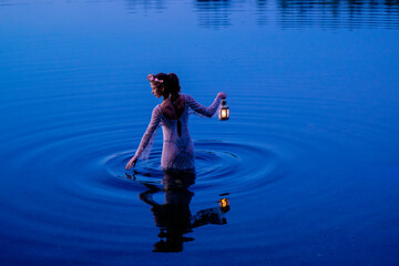 Woman in patterned dress holds lantern standing in serene blue lake at dusk. WA, USA