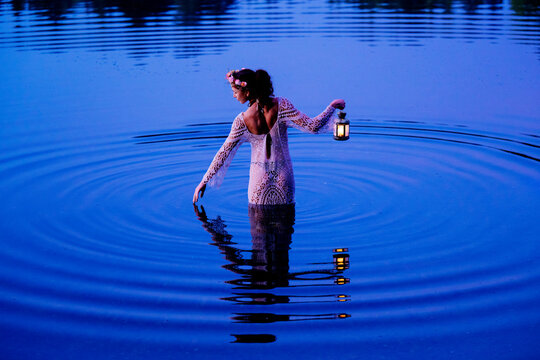 Woman in lace dress wades in calm lake at dusk, holding a glowing lantern. WA, USA