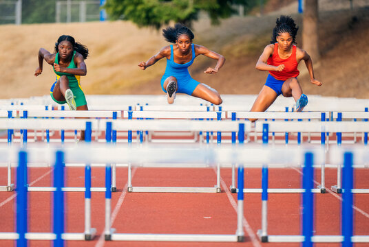 Three athletes sprint and leap over hurdles on a track in a competitive race. USA