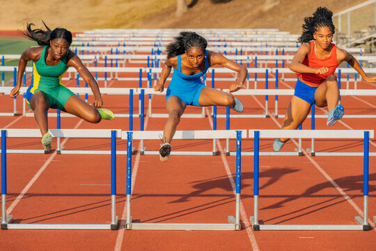 Three athletes hurdle on a red track under sunny skies, displaying strength and determination. USA