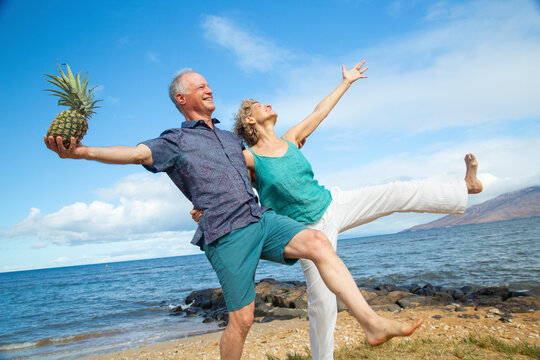 Senior couple balancing on one leg by the beach, sharing a playful moment with a pineapple. Kepaniwai Park, Wailuku, Hawaii - Powered by Adobe