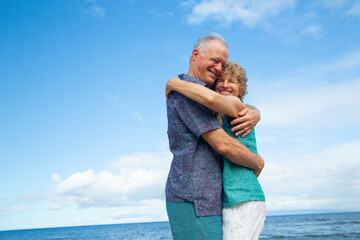 Elderly couple embracing on a beach under a bright blue sky with ocean in background. Kepaniwai Park, Wailuku, Hawaii