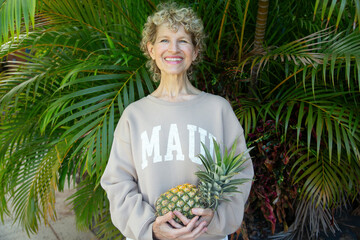 Smiling woman holding a pineapple in front of lush green tropical plants. Kepaniwai Park, Wailuku, Hawaii
