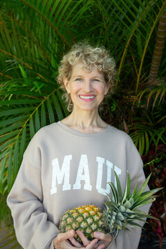 Smiling woman holding a pineapple in front of lush green palm leaves. Kepaniwai Park, Wailuku, Hawaii
