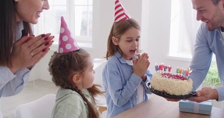 Smiling family wearing festive hats joyfully celebrating their son birthday at home. Parents and sister happily watching as excited boy blowing out candles on his cake while they clapping together.