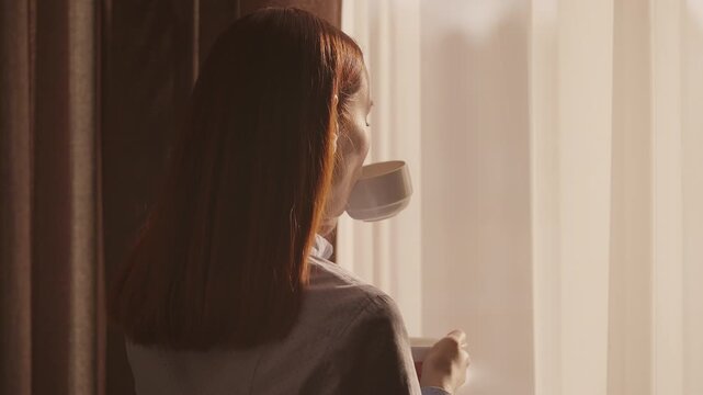 Happy red-haired woman drinking morning coffee by window, resting in hotel room