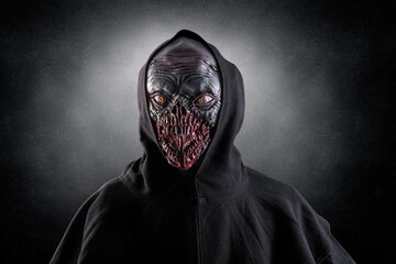 Portrait of a monster with sharp fangs with hooded cape over dark foggy background. 