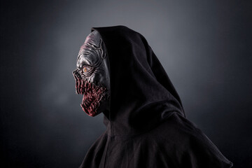 Portrait of a monster with sharp fangs with hooded cape over dark foggy background. 
