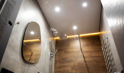 Modern bathroom with wood tiles, mirror and stretch ceiling with LED lighting. Chrome shower and heated towel rail, modern renovation