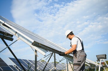 Portrait of Young indian man technician wearing white hard hat near solar panels against blue sky....