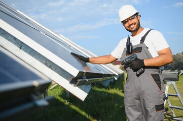Portrait of Young indian man technician wearing white hard hat near solar panels against blue sky....