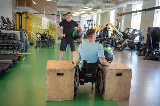 Determined man in wheelchair do guided therapy sports, lifting and balancing heavy medical ball under personal trainer supervision. Male focused on shoulder stability core activation, spinal trauma