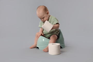 Cute little baby sitting on potty and rolls of toilet paper against light grey background
