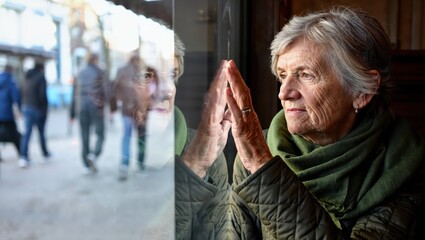 Thoughtful elderly woman looking out window with reflection and nostalgia.