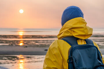 Luc-Sur-Mer, France - 08 10 2025: A woman wearing a yellow raincoat and a hat on the jetty looking the sunset with the sea, the beach and the sky behind