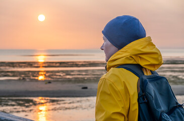Luc-Sur-Mer, France - 08 10 2025: A woman wearing a yellow raincoat and a hat on the jetty looking the sunset with the sea, the beach and the sky behind