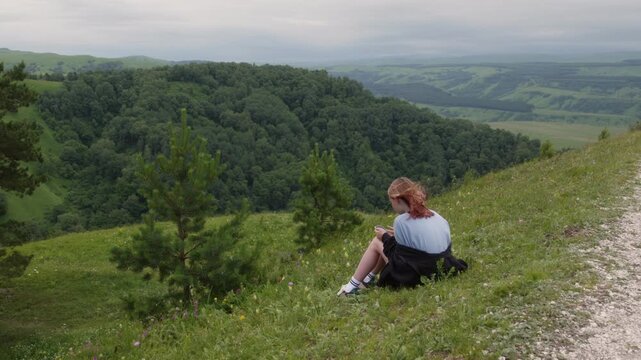 Resting on a lush green hillside, a young woman hiker pauses to jot down notes, surrounded by the tranquility of nature and a picturesque view of rolling hills and valleys