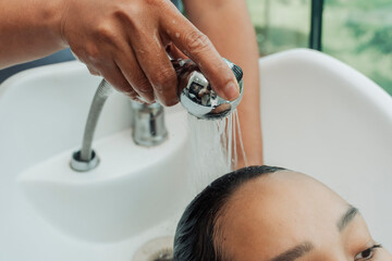 Water flows directly onto the scalp of a Southeast Asian woman at a salon sink, preparing for...