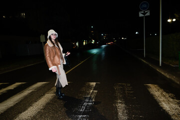 Woman crossing a quiet street at night wearing a stylish coat and boots, illuminated by distant...
