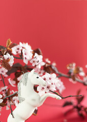A white horse is standing on a red background with pink flowers. The horse is looking up at the camera, and the flowers are in full bloom. Concept of beauty and serenity, as the horse