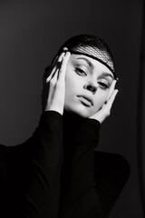 Stylish young woman posing with hands on face, wearing a black outfit and fishnet headpiece....