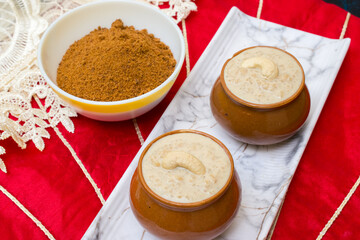 Jaggery rice kheer or pudding served in a small clay pot, topped with cashew nuts and placed on a white tray with jaggery powder in a bowl, against a vibrant red festive background. 