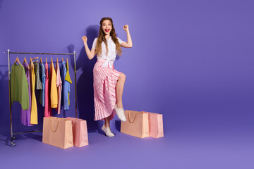 Cheerful young woman shopping with colorful wardrobe and bags on purple background, expressing joy...