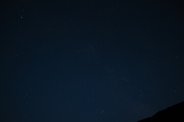 Naklejka premium Starry night sky above mountain silhouette, with faint meteors and constellations visible in deep blue. View of the Milky Way from the northern hemisphere.