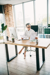 Confident mature businesswoman analyzing documents at a modern office desk with computer and...