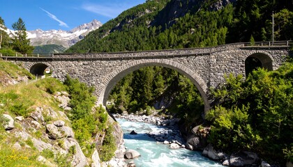 Fototapeta premium A grand stone arch bridge spans a rushing river, nestled amongst lush green alpine scenery and snow-capped peaks.