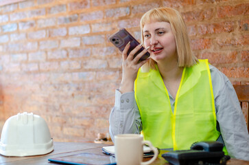 Smiling young blonde industrial employee wearing safety gear sending an audio message with her...
