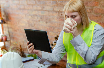 Young blonde industrial employee wearing safety gear using digital tablet while drinking coffee at office or home office