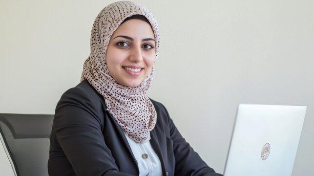 A happy and professional Muslim businesswoman wearing a hijab and formal attire, smiling while working on her laptop in a modern office.