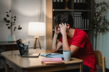 Young man struggling with a headache while working late on a laptop at home, feeling overwhelmed by...
