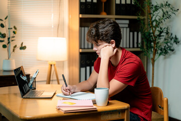 Teenager focused on writing in a notebook while utilizing a laptop for online learning at home,...