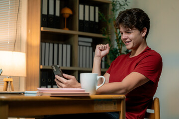 Young man expressing excitement while using smartphone at his desk at home, celebrating a positive online result