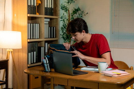 Focused student searching bag contents while sitting at desk, laptop nearby, studying late at home with coffee cup - Powered by Adobe