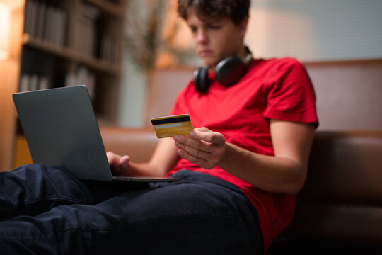 Young person completing online transaction, holding credit card while sitting on living room sofa with laptop open