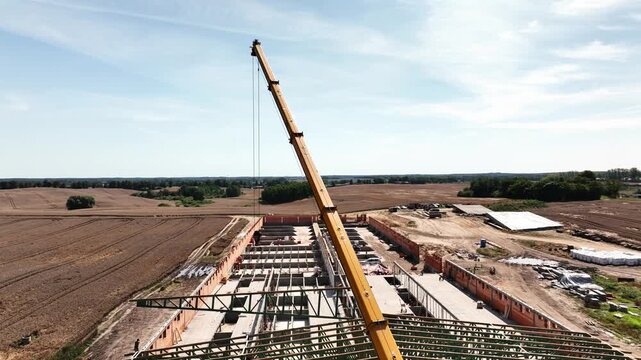 Smooth drone orbit around mobile telescopic crane lowering wooden roof truss onto modern pig farm construction site. Agriculture, engineering and rural progress concept