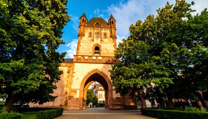 A substantial ancient gateway with a tall tower, adorned with intricate stonework, stands under a partly cloudy sky, bathed in warm sunlight.