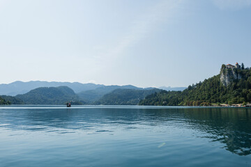 Scenic view of Lake Bled with calm blue water, surrounded by green forest and mountains on a clear summer day in Slovenia. Old church on the island and castle on the rock