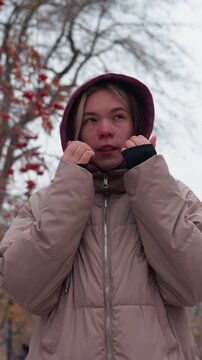 Woman bundled in warm winter clothing adjusts her hoodie while walking through a snowy park, she holds her hands near her face to keep warm, surrounded by bare trees with red berries
