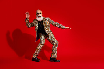 Stylish elderly man in leopard print suit and sunglass posing on vibrant red background with a confident and funky attitude.