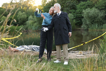 Forensic expertise. Detectives working at crime scene with dead man's body near lake outdoors