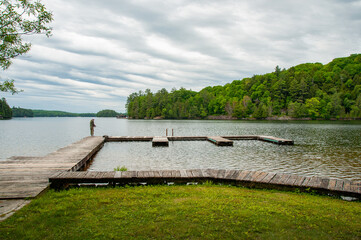 A man fishing at the end of a dock on a beautiful forested lake during a cloudy day in the summer.
