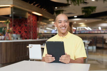 Fototapeta premium Middle-aged bald Hispanic man using tablet computer while dining at food court restaurant
