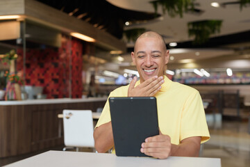 Middle-aged bald Hispanic man using tablet computer while dining at food court restaurant