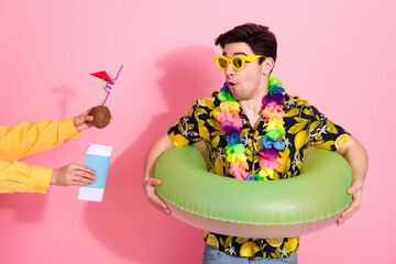 Young man with tropical attire wearing sunglass and inflatables, receiving a drink against pink...